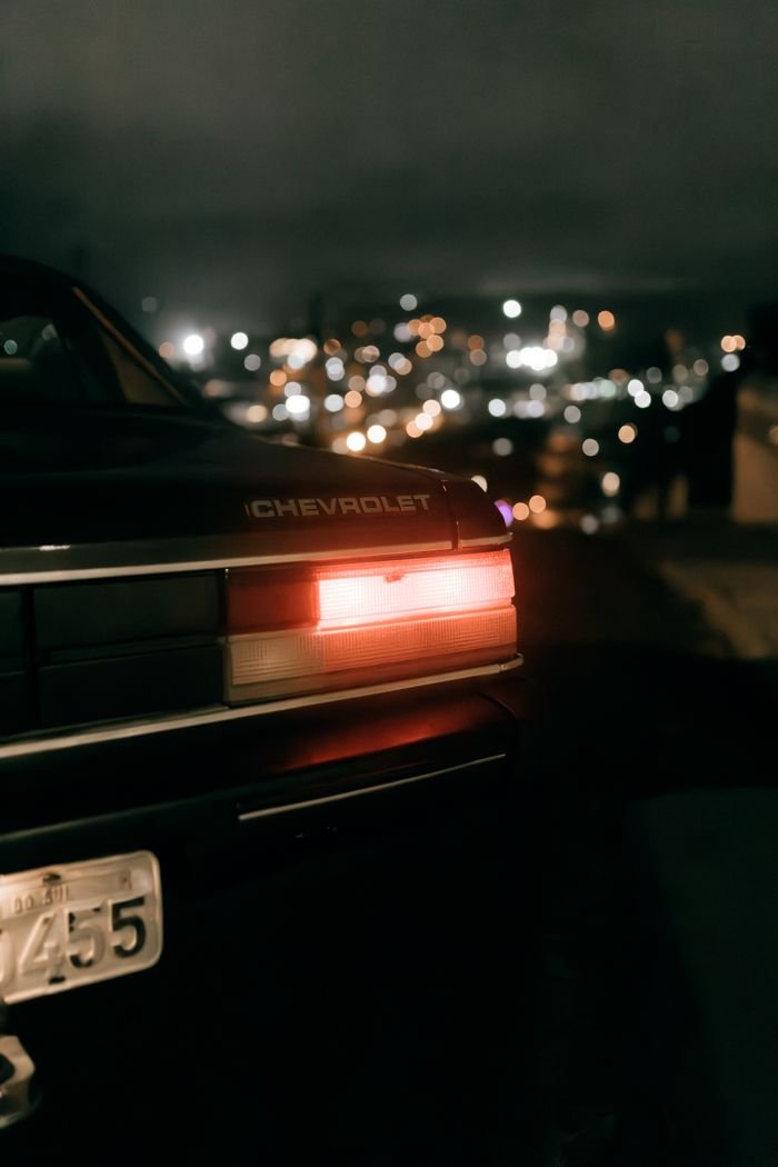 Vintage Chevrolet car with tail lights on, overlooking city lights at night, creating a moody atmosphere.